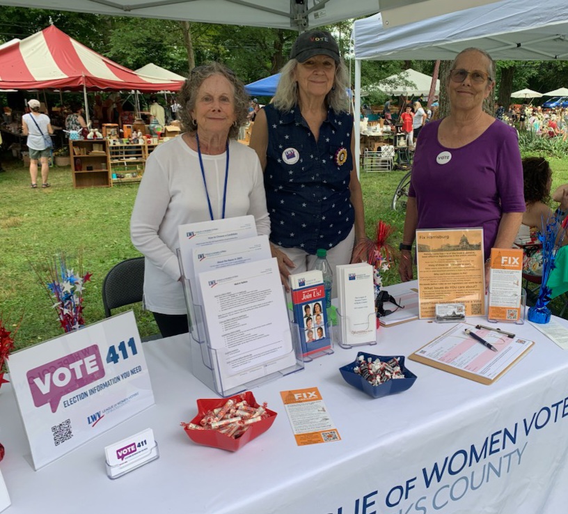 3 older, caucasian female, league members standing at a community event table.  table has materials about the league