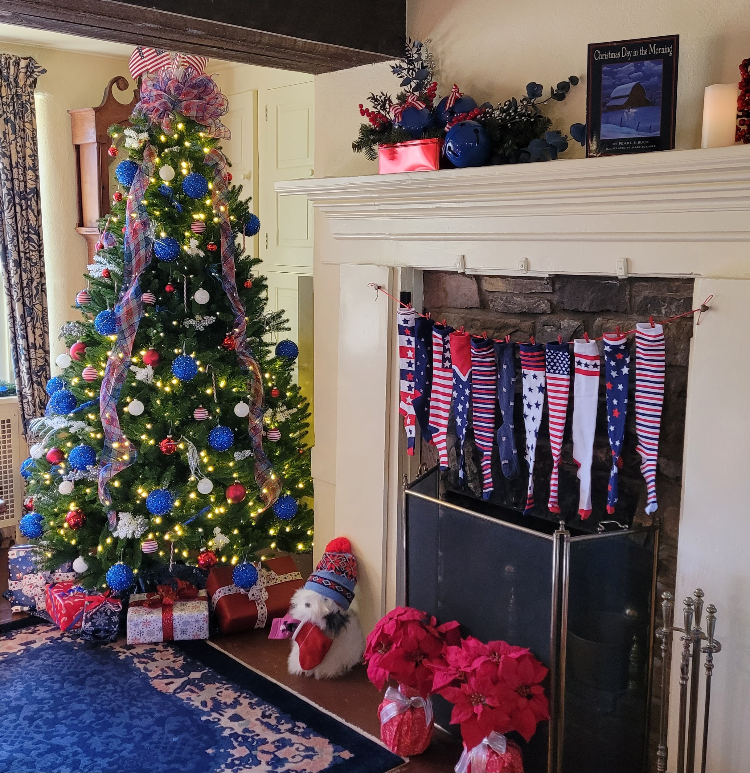 Color photograph of a decorated Christmas tree in front of a fireplace.  Fireplace mantel has red, white and blue patterned holiday stockings