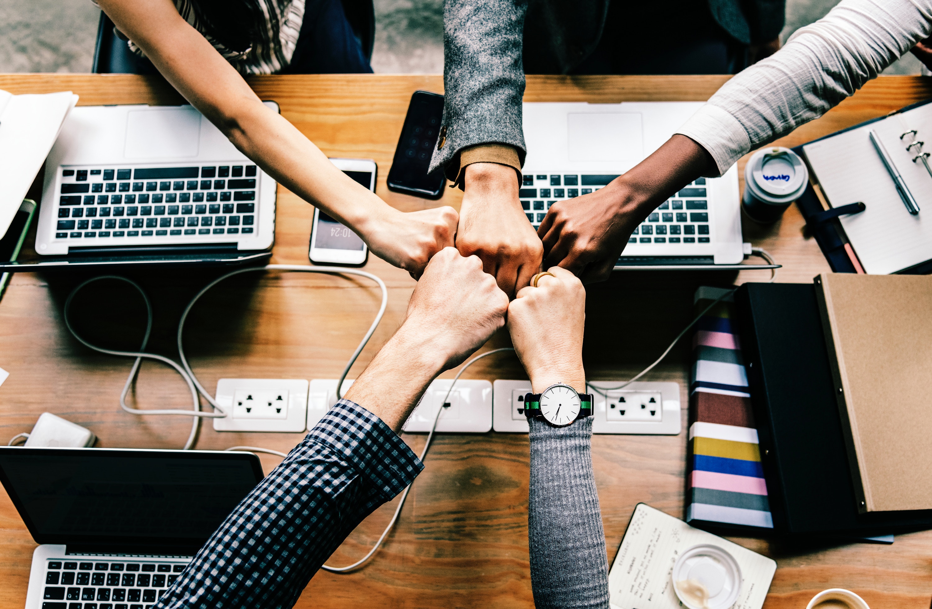 A top-down image of five fists of varying sizes and skin tones being held together over a table with computers, wires, and smartphones on it. The people appear to be wearing attire for working in an office. A top-down image of five fists of varying sizes and skin tones being held together over a table with computers, wires, and smartphones on it. The people appear to be wearing attire for working in an office.