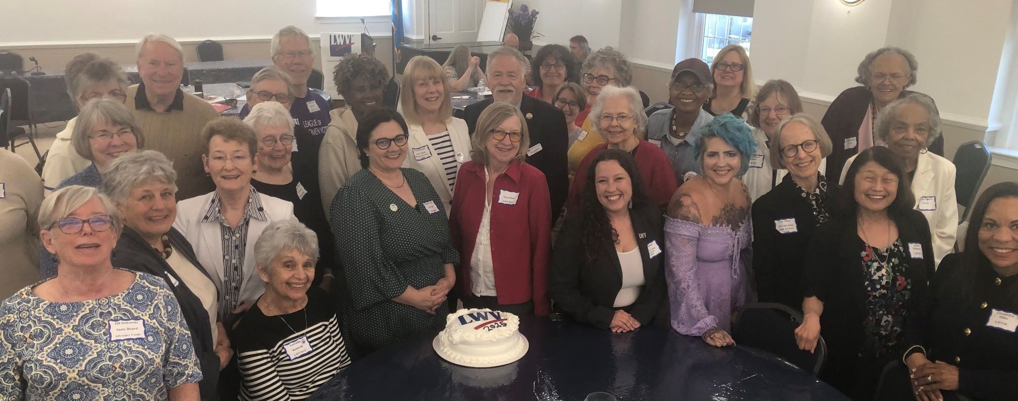 30 plus convention attendees are shown gathered around a table with an LWV cake, lookup up at the photographer