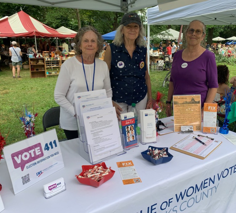 3 older, caucasian female, league members standing at a community event table. table has materials about the league