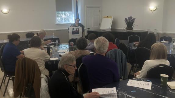 TIffany Lydon is shown at the podium, with attendees seated at round tables in foreground