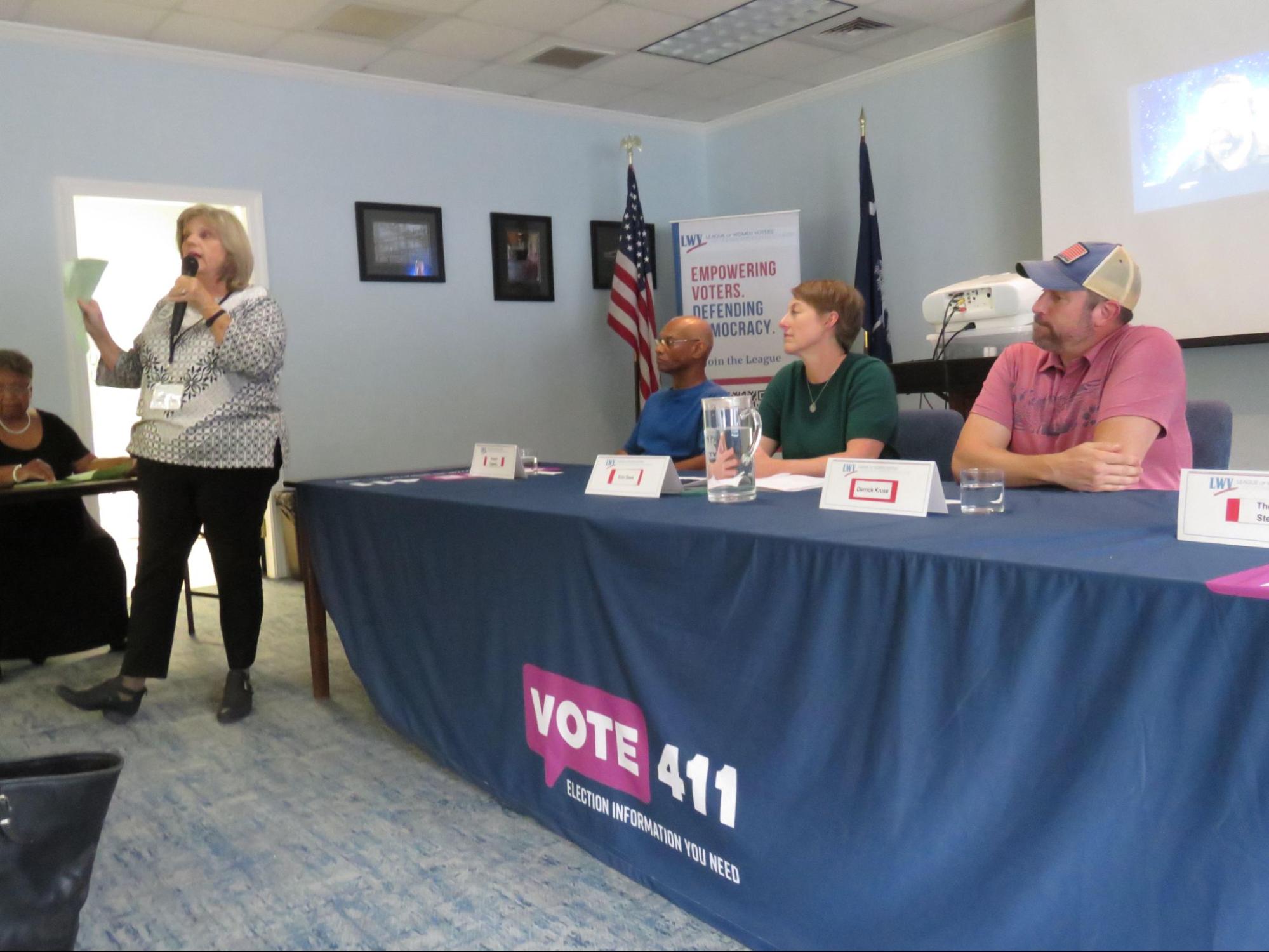 A woman talks in a microphone while two people seated at tables listen.