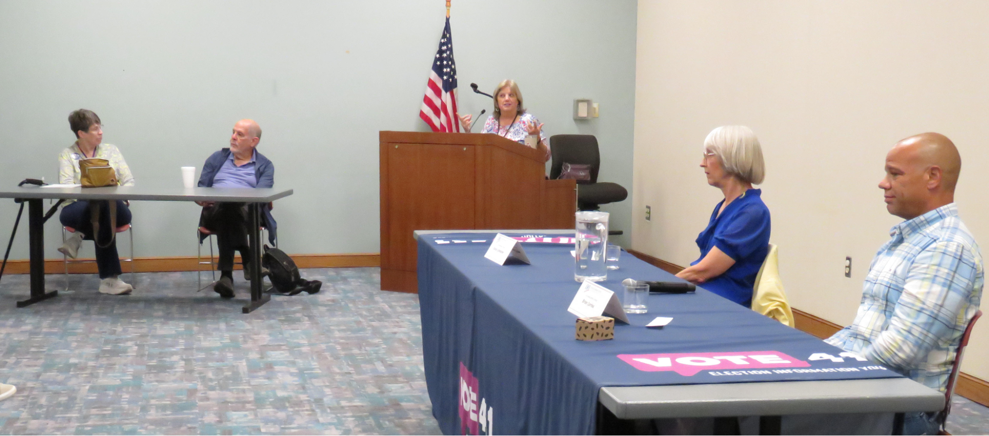 Four people seated at tables listen to a woman at a lectern at a candidate forum