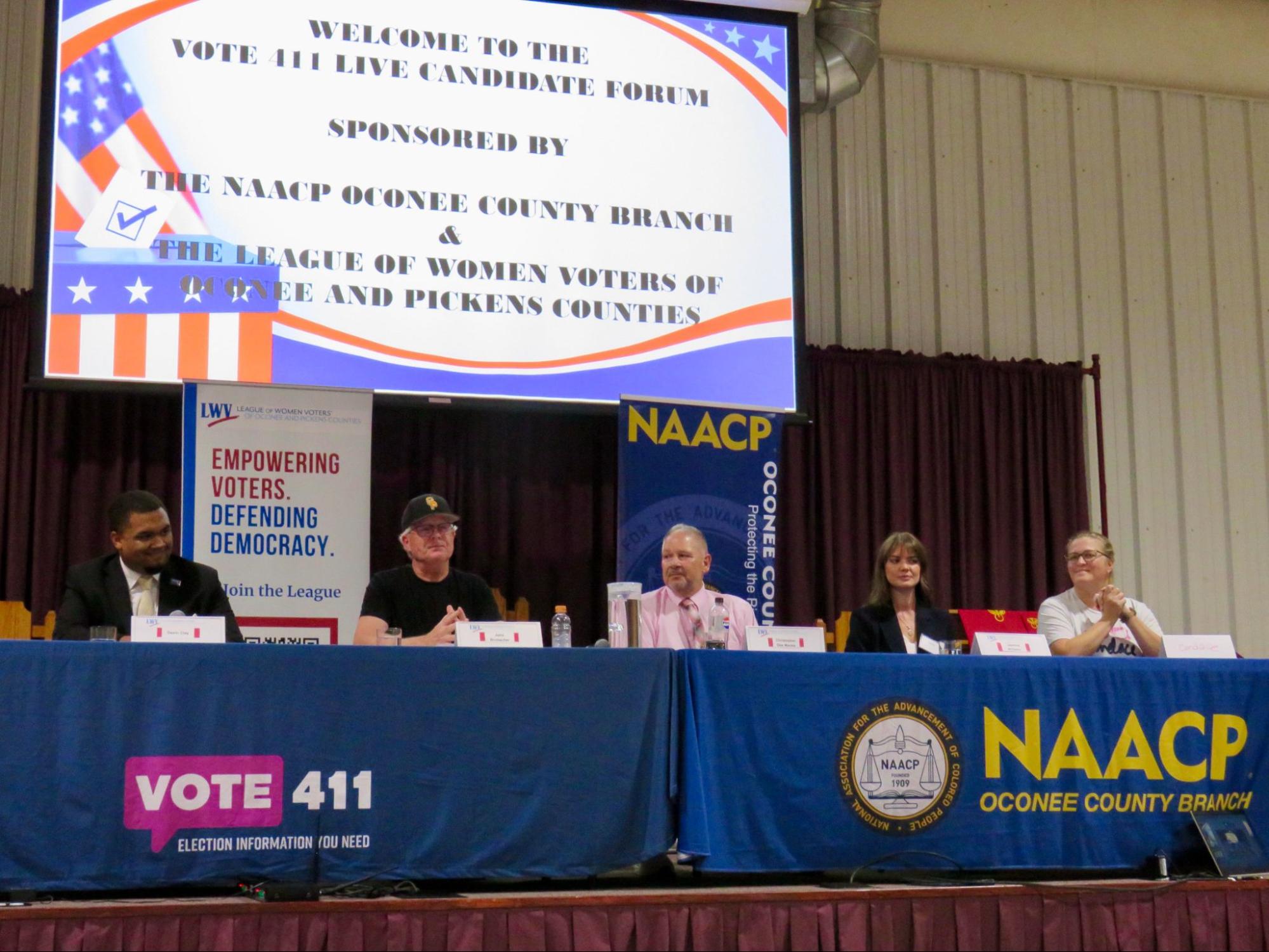 Five people sit behind tables with Vote411.org and NAACP table drapes. Behind them is a screen that welcomes people to the candidate forum.