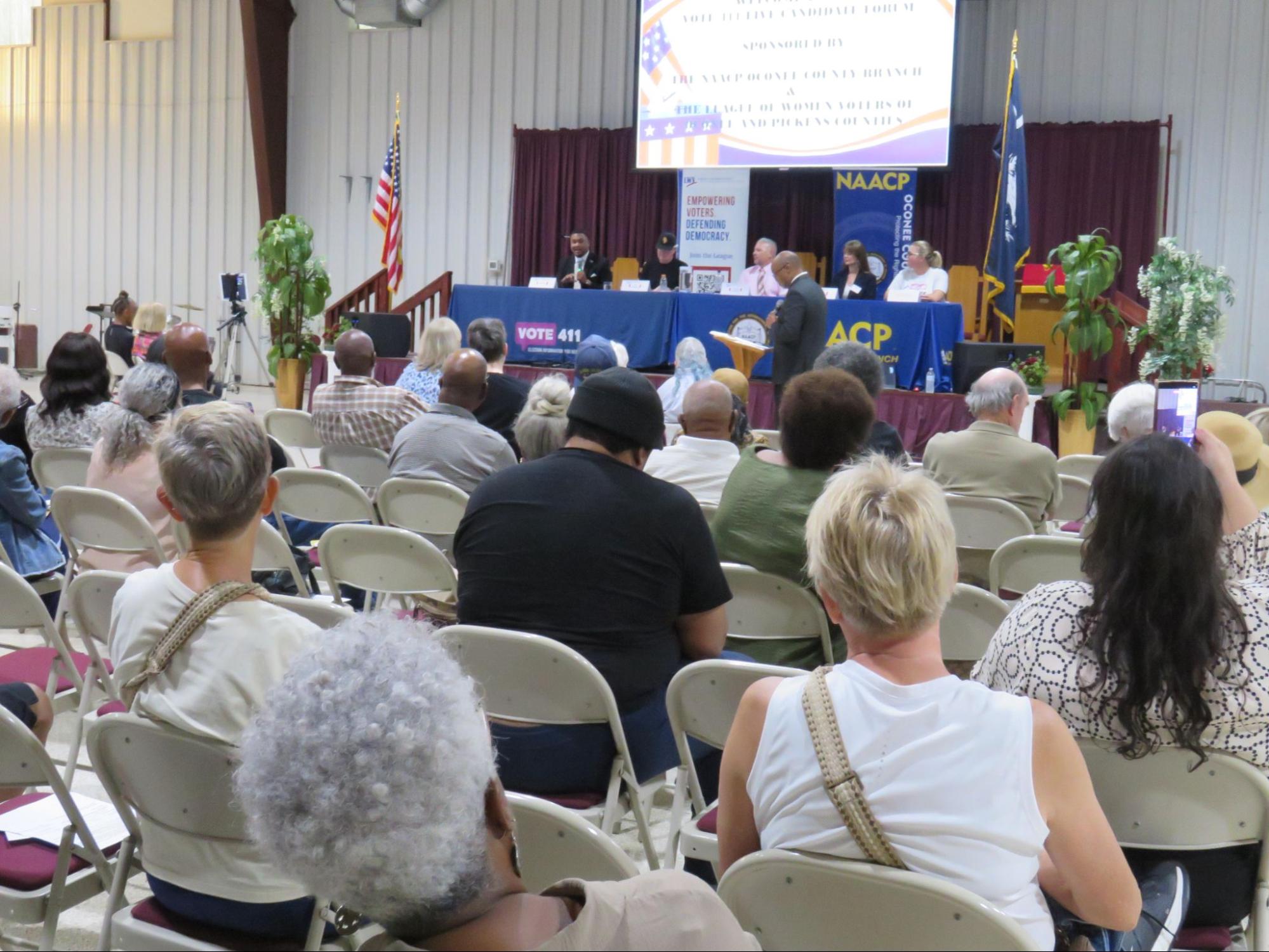 People sitting in chairs watching a five people behind tables at a candidate forum