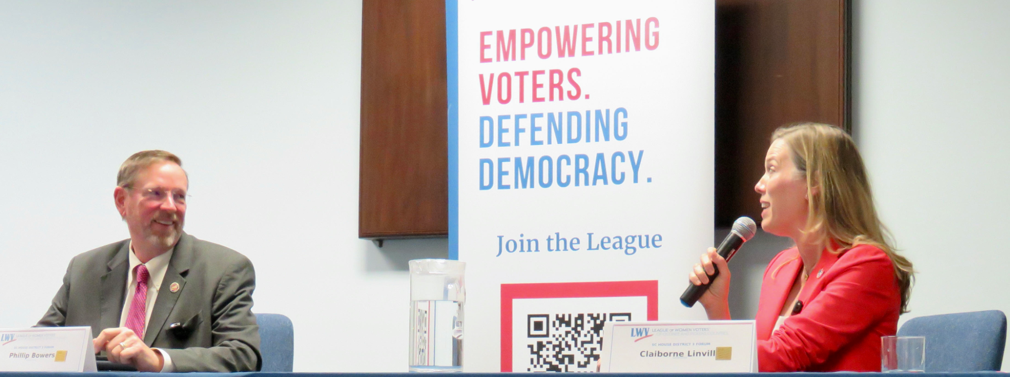 A man listens to a woman holding a microphone. They both sit at a table with a sign behind them that reads "Empowering Voters. Defending democracy."