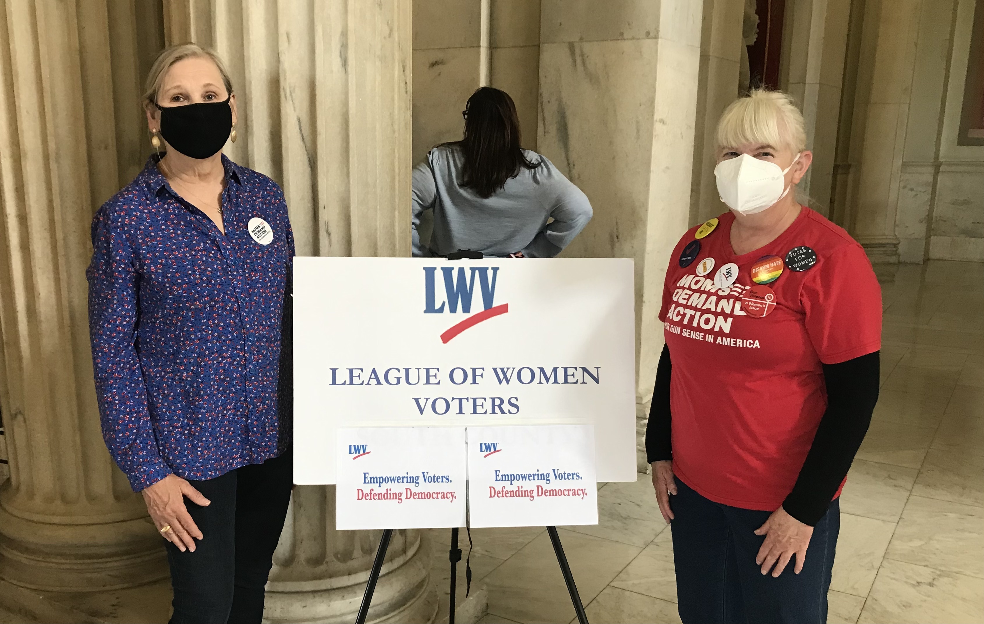 Two members of the League of Women Voters RI South County Chapter standing in front of a League sign on an easel.