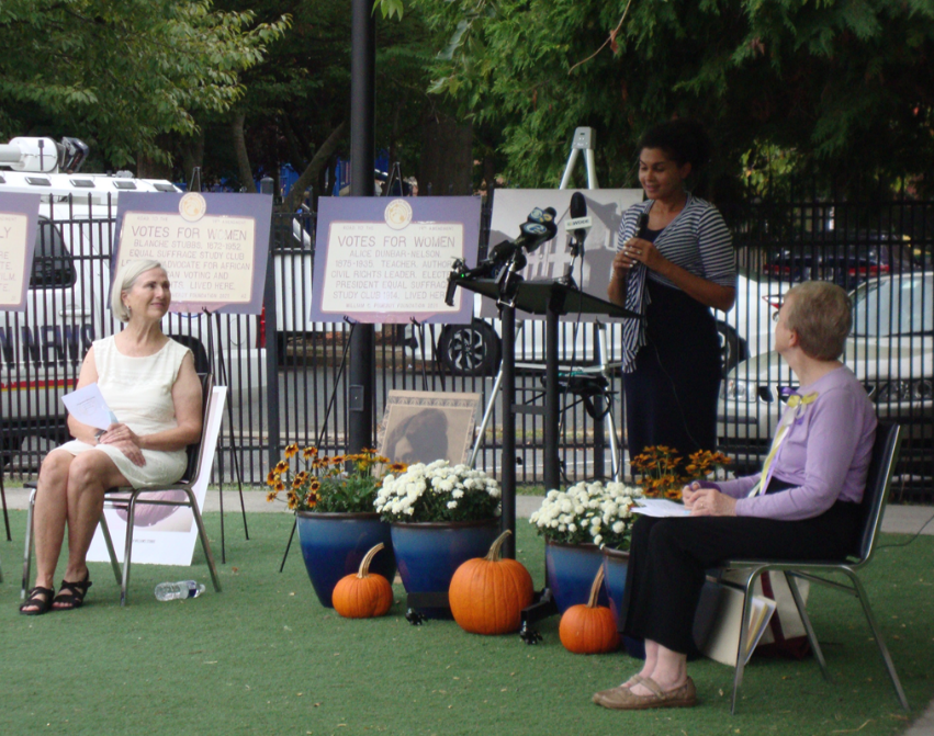 photo of standing and seated women presenters at outdoor ceremony with green grass, pumpkins, fall flowers and signboards arranged on easels