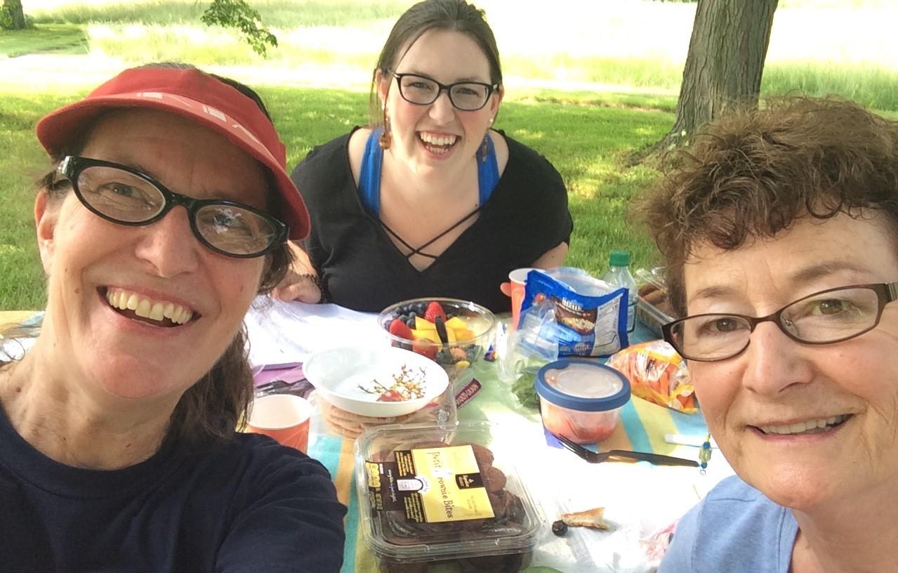 Three women at a picnic table in a grassy area, photo taken selfie-style