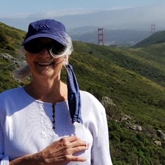 Head and shoulders photo of white woman with gray hair wearing a white shirt and blue ball cap and sunglasses with the Golden Gate Bridge in the background