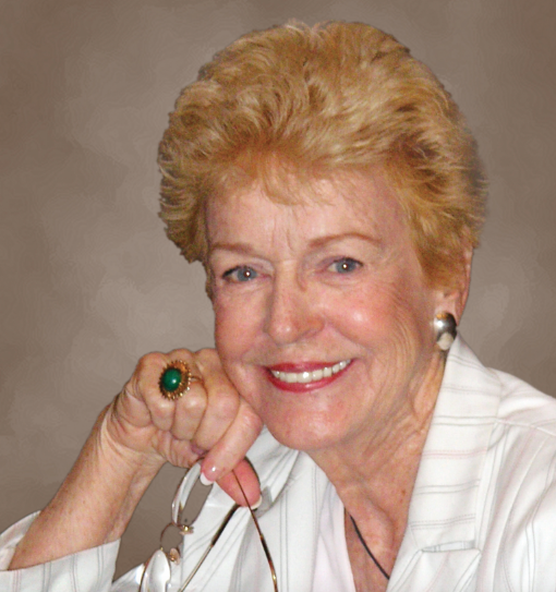 Headshot of older woman with red/blonde hair, white shirt and green ring. The backdrop is gray.