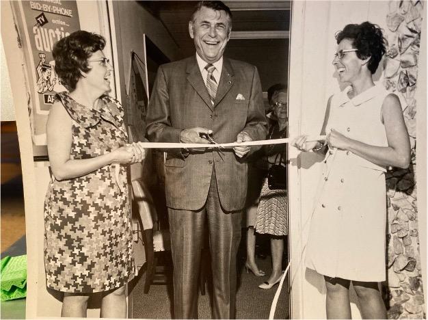 Sepia image from 1971 of two women holding a ribbon and a man in a suit cutting the ribbon