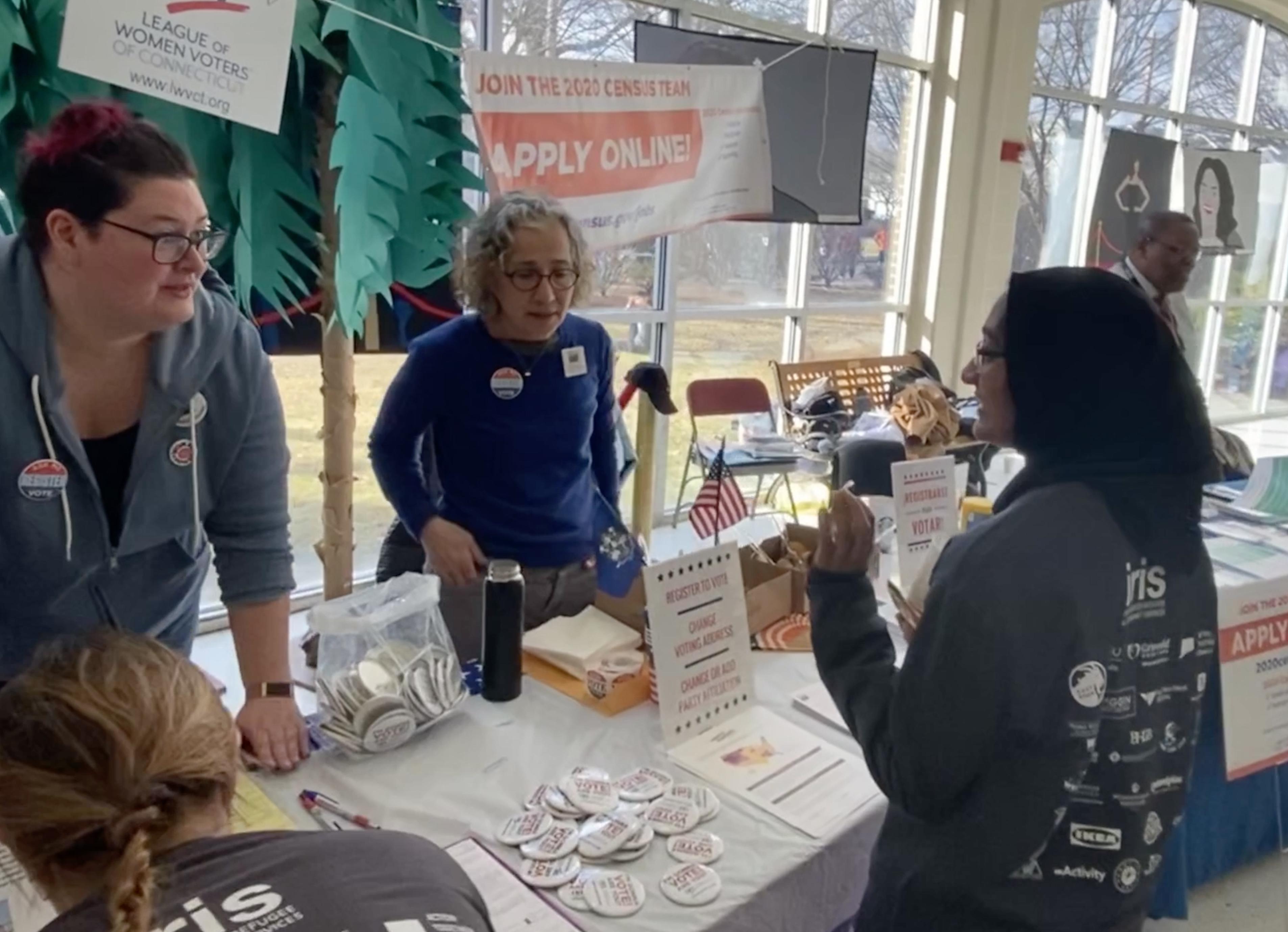 Two women at a table with lots of materials on it speaking to a third woman about registering to vote