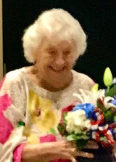 A smiling woman with short white hair and a colorful top accepts a bouquet of colorful flowers