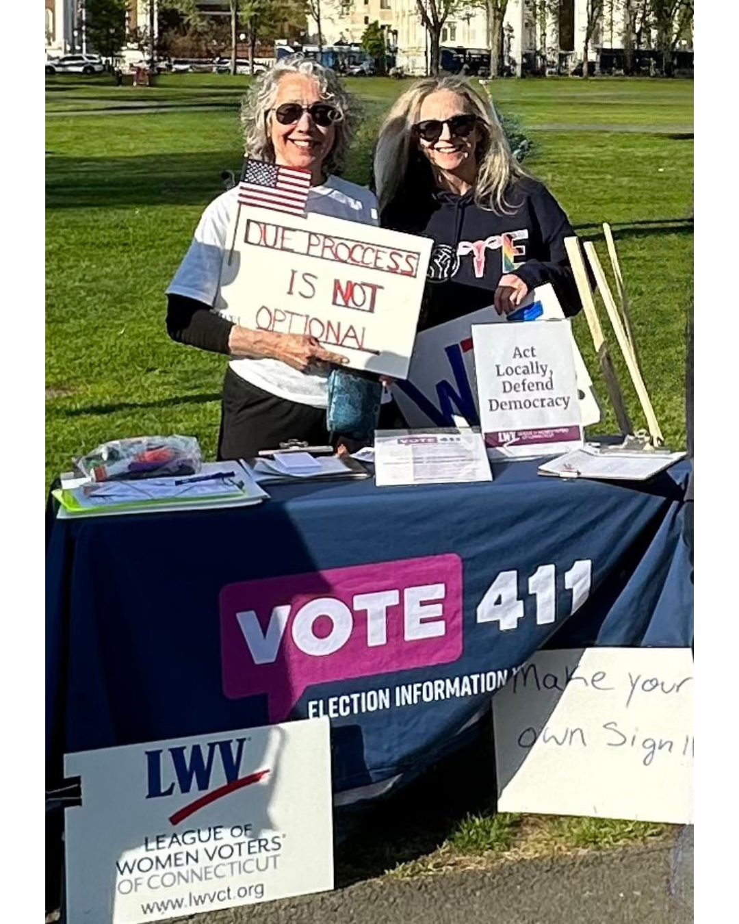 Laura Smits and Patricia Rossi behind a VOTE411 table holding signs that say "due process is not optional" and "Act Locally, Defend Democracy"