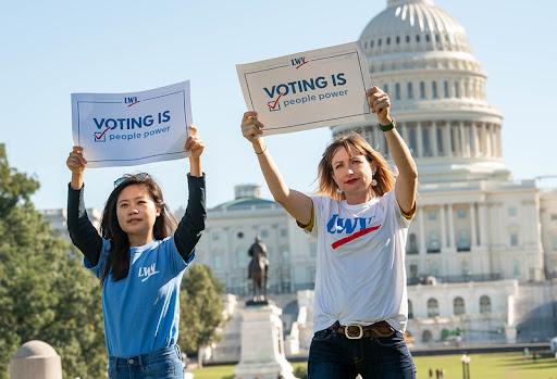 Two women with LWV t-shirts standing in front of the US capitol building holding signs that say Voting Is People Power
