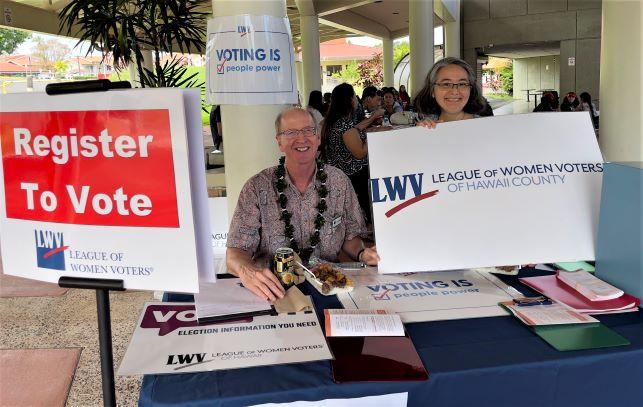 Chuck Greenfield sitting at voter registration table and Michele Mitsumori standing holding a League of Women Voters sign, both smiling