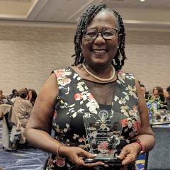 Smiling woman with dark skin, dark hair, glasses and a floral top holding an award