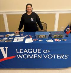 A brunette woman shown standing behind a League of Women Voters information table