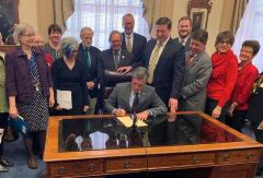 Photo shows Delaware Governor Carney at a large glass desk signing the National Popular Vote Interstate Compact Bill in January 2020, surrounded by supporters of the bill.