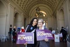 Older and younger women pose together holding a sign each that reads: WOMEN POWER THE VOTE