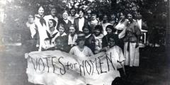 Group photo of Sufferagettes holding Votes For Women sign