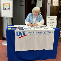 Julia is shown doing paperwork at a table featuring League of Women Voters information.