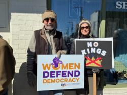 Rally participants holding signs, one says "Women Defend Democracy" and the other says "No Kings"