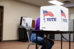 person sitting at a table behind a screen labeled VOTE