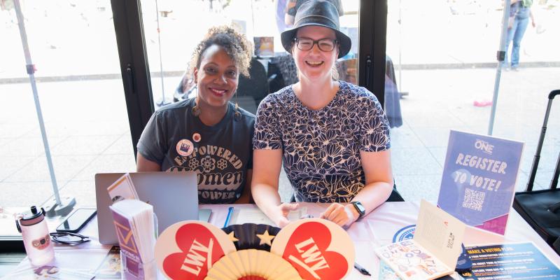 Two smiling people sit behind a table at a civic engagement event