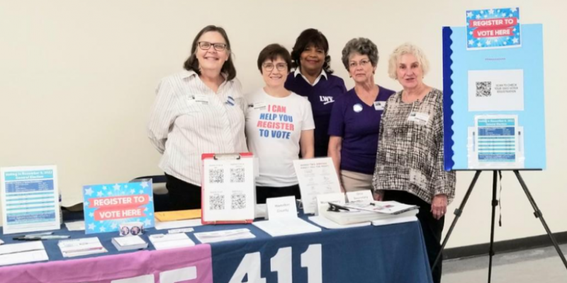 5 women smile at the camera.  They are standing behind a table filled with paperwork and an easel with informational posters.