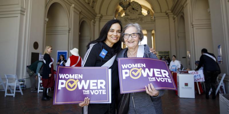 Older and younger women pose together holding a sign each that reads: WOMEN POWER THE VOTE