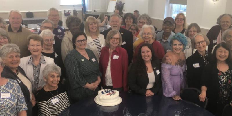30 plus League members in attendance are gathered around a table and an LWV cake, looking up at photographer