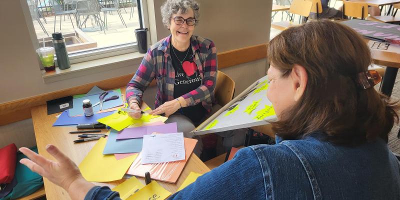 Two women are shown in the midst of creative chaos with colored paper, scissors and a white board