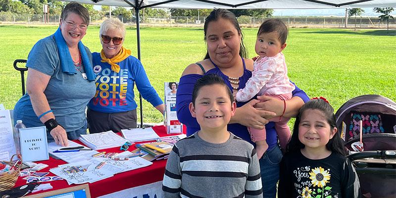 A Mom with infant and two children under the umbrella with two woman at the League of Women Voter's table