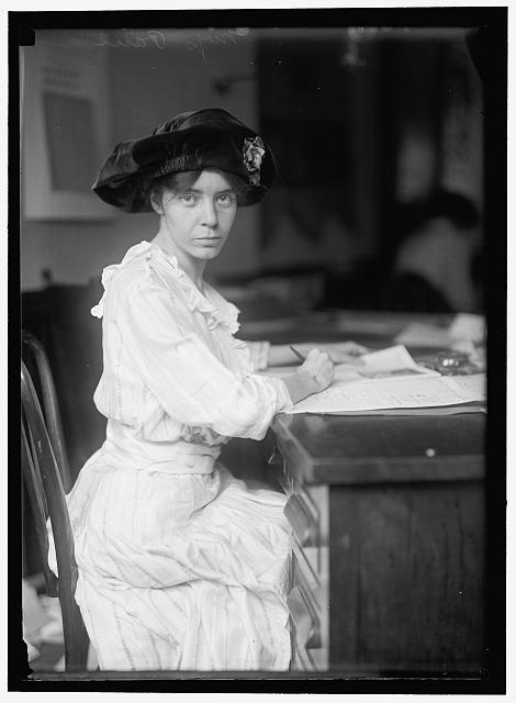 Vintage photo - Woman with dark hair and hat sits at desk and looks to the camera